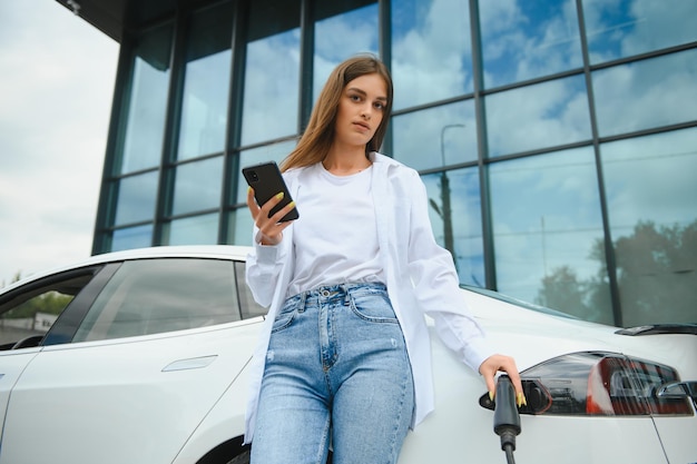 Phone in hands Woman on the electric cars charge station at daytime Brand new vehicle