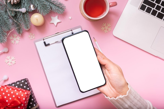 Phone mock up in female hand with Cristmas New Year work table decoration top view flat lay on pink background