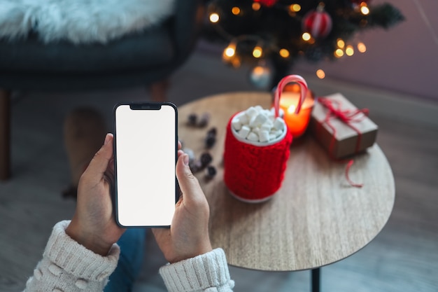 Макет телефона, руки с пустым экраном для текста на фоне новогоднего декора.