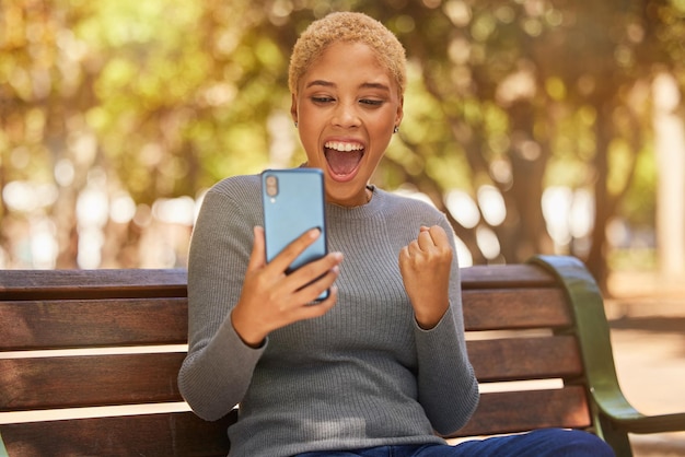 Phone success and woman excited about trading on the internet while on a bench in the park Young happy and girl with wow face for communication on a mobile app celebration and winning in nature