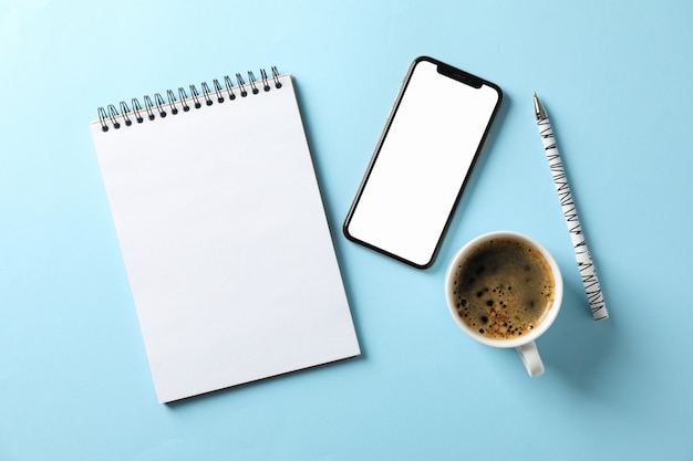 Phone with empty screen, copybook, pen and cup of coffee on blue background, top view