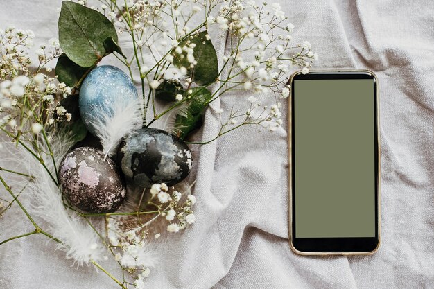 Foto telefono con schermo vuoto ed eleganti uova di pasqua piatte su un tavolo rustico con fiori primaverili bianchi uova di pasqua colorate tinte naturali su tessuto rurale con smartphone spazio per il testo