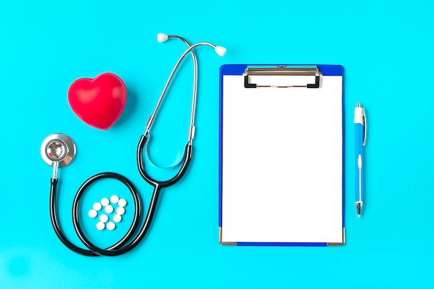 Phonendoscope, heart, tablet with white paper, pen, medicine on blue background