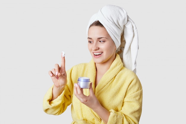 Phot of joyful healthy female holds container with cream, satisfied with its good effect, dressed in casual clothes, bathrobe and towel, has beauty treatments at home, isolated over white wall