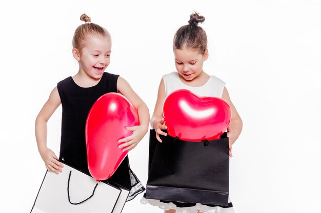 Photo of 2 little girls with elegant dresses hold large bags with heart-shaped balloons inside