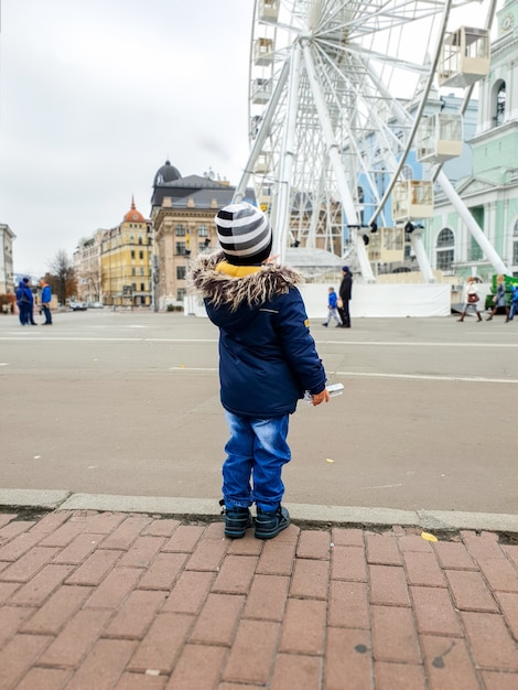 Фотография 3-летнего мальчика, смотрящего на высокое колесо обозрения на городской улице