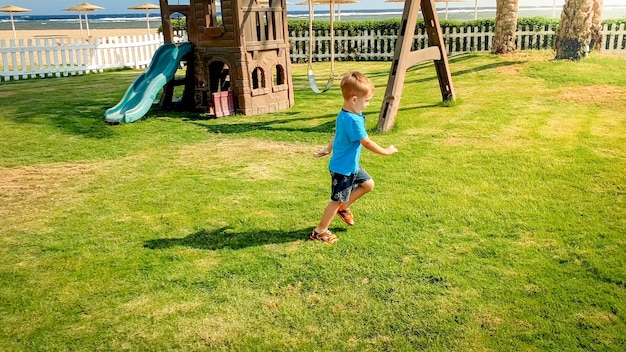 Photo of 3 years old toddler boy running on big palyground with beatiful grass lawn at park