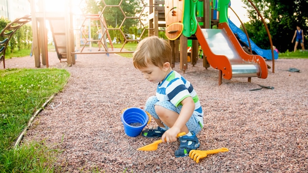 Photo of adorable 3 years old little boy sitting on the playground and digging sand with small plastic shovel and bucket