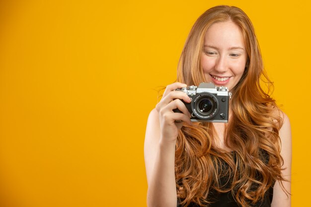 レトロなビンテージカメラを押しながら黄色の壁コピースペースで分離された写真を撮る黒い服を着て愛らしい赤毛の女性の写真。レトロなビンテージ写真カメラを保持します。