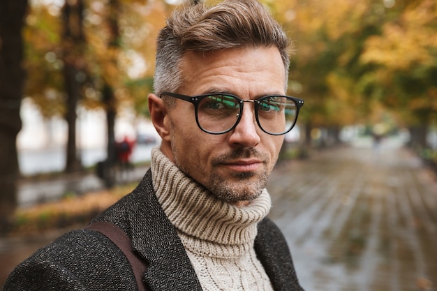 Photo of adult man 30s wearing eyeglasses, while walking outdoor through autumn park