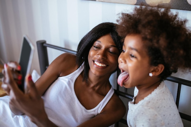 Photo of african american mother and daughter taking a picture with mobile phone in the bed