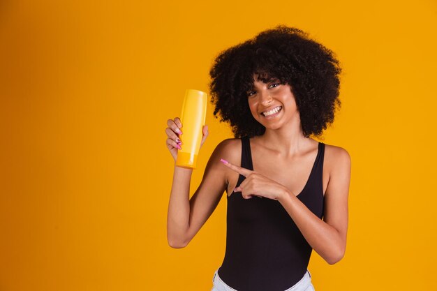 Photo of afro natural brunette holds gel shampoo from bottle looking at camera over yellow background