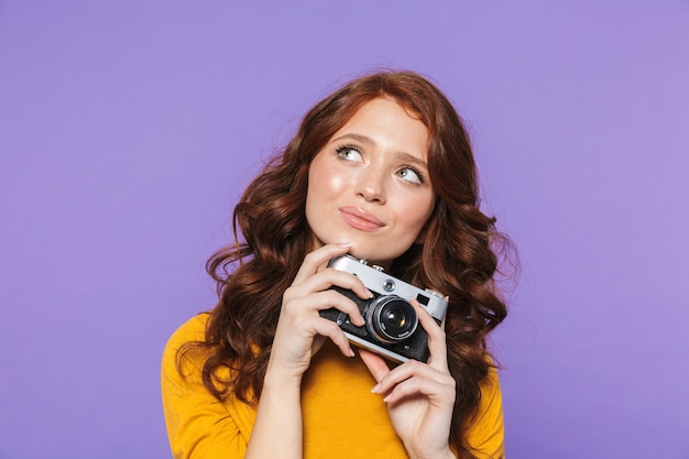 Foto della donna dai capelli rossi seducente che indossa vestiti gialli che tiene la retro macchina fotografica d'annata e che cattura l'immagine sopra la porpora