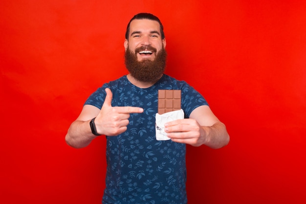 Photo of amazed bearded man pointing at bar of chocolate over red background