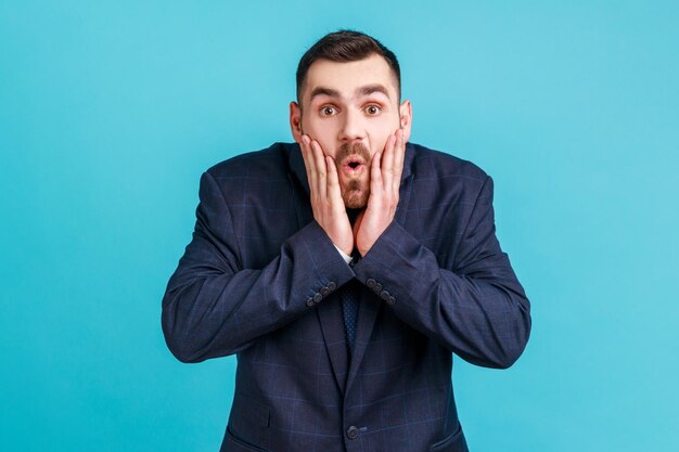 Photo of amazed bearded young adult man in official style suit\
looks at camera with open mouth raising palms being impressed of\
shocking news indoor studio shot isolated on blue background