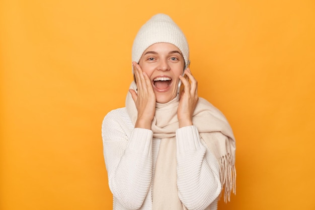 Photo of amazed happy excited young adult woman wearing warm sweater cap and scarf posing isolated over yellow background talking on smart phone hearing good news