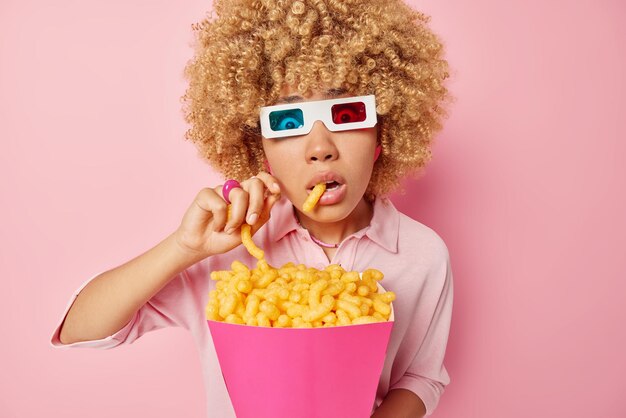 Photo of amazed woman holds breath as watches horror film in\
cinema eats delicious corn sticks from paper bucket wears 3d\
eyeglasses casual shirt isolated over pink background entertainment\
concept