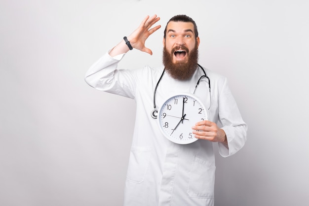 Foto di un giovane dottore stupito che tiene in mano l'orologio da parete e fa gesti