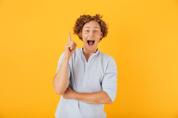 Photo of amazed young man  screaming and pointing finger upward at copyspace, isolated over yellow background