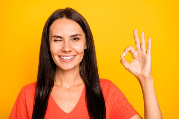 Foto foto di sorprendente giovane donna ammiccante occhio che mostra okey simbolo esprimere accordo consigliando prodotto indossare casual maglietta arancione isolato giallo parete di colore