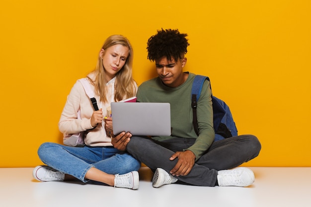 Photo of american and european students 16-18 using silver laptop while sitting on floor with legs crossed, isolated over yellow background