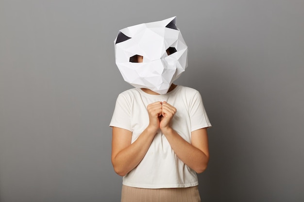 Photo of anonymous cute charming woman wearing cat paper mask and white Tshirt posing isolated over gray background standing looking at camera keeps hands together