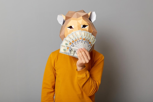 Photo of anonymous man wearing lion mask and orange sweatshirt isolated over gray background standing covering half of face with with dollar banknotes