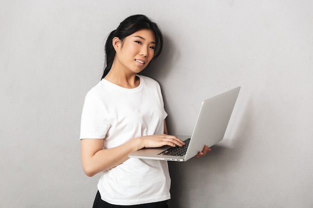 Foto di bella giovane donna asiatica che posa isolata sopra la parete grigia facendo uso del computer portatile.