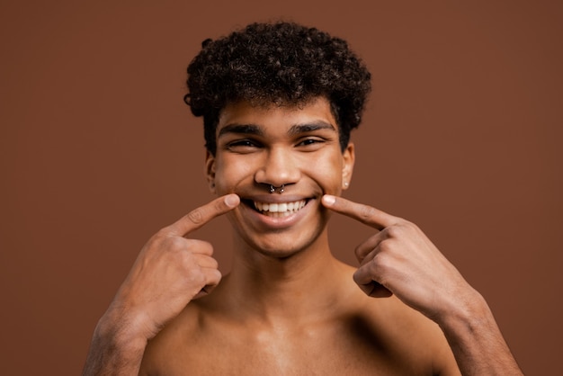 Photo of attractive black man with piercing show how wide his smile is. Naked torso, isolated brown color background.