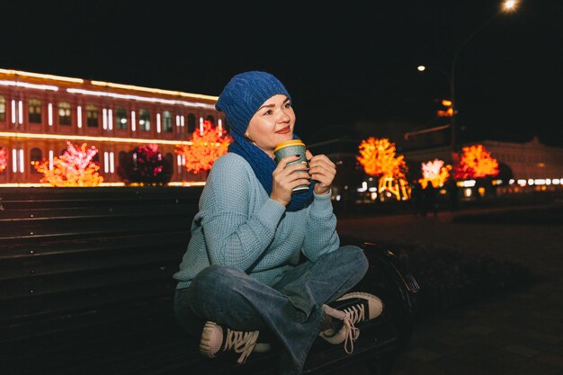 Photo of attractive cheerful woman sitting on a bench in cold snowy weather, smiling, drinking coffee outdoors