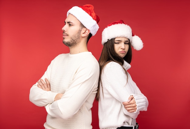 Photo of attractive couple in Christmas hats, stands back to back and looks resentful and desperate, isolated over red background.