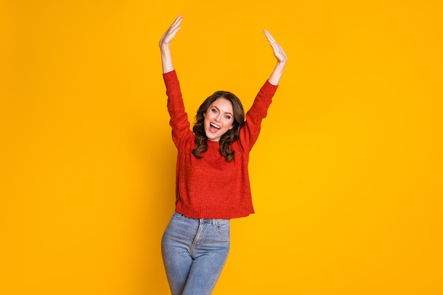 Photo of attractive curly funny lady good mood raise arms cheerful person rejoicing overjoyed wear red knitted sweater blue jeans isolated bright yellow color background