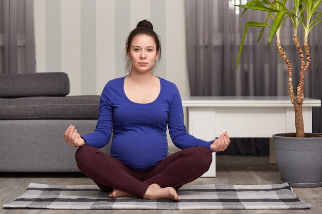 Photo of attractive dark haired expectant mother sits in lotus pose