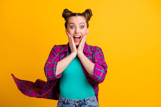 Photo of attractive excited crazy teen lady two funny buns good mood arms on cheeks overjoyed listen amazing news wear casual plaid shirt isolated yellow bright color background