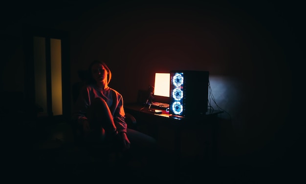 빨간 불빛 아래 컴퓨터 근처의 의자에 앉아 있는 매력적인 소녀의 사진 컴퓨터에서 야간 작업