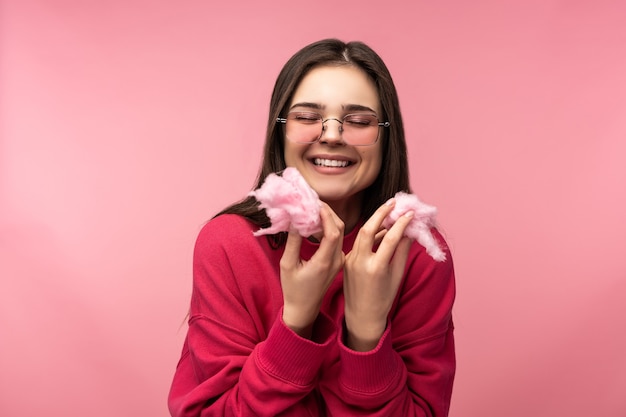 Photo of attractive lady in glasses plays with cotton candy, has fun. Wears casual pink sweater white pants isolated pink color background.