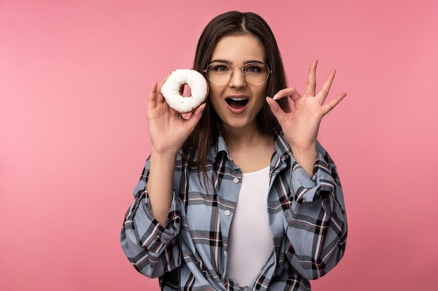 Photo photo of attractive lady holds donuts smile, happy shows ok, wear casual checkered shirt white undershirt isolated pink color background.