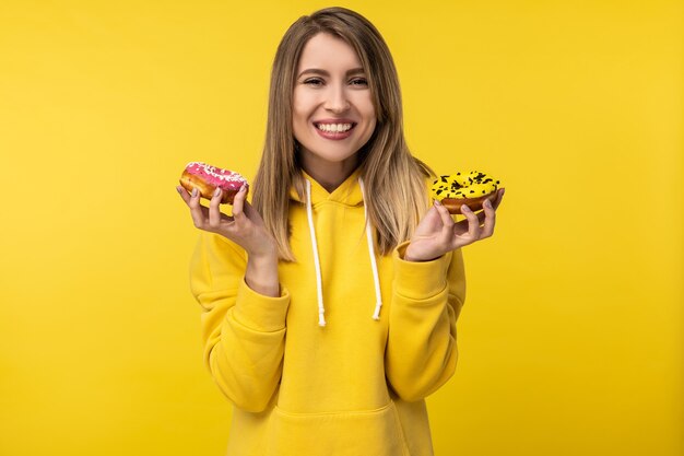 Photo of attractive lady holds donuts with smile. Wears casual yellow hoody, isolated yellow color background.