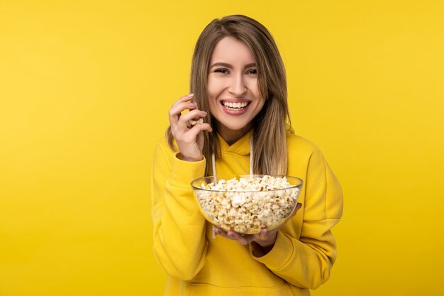 Photo of attractive lady holds plate of popcorn, with smile. Wears casual yellow hoody, isolated yellow color background.