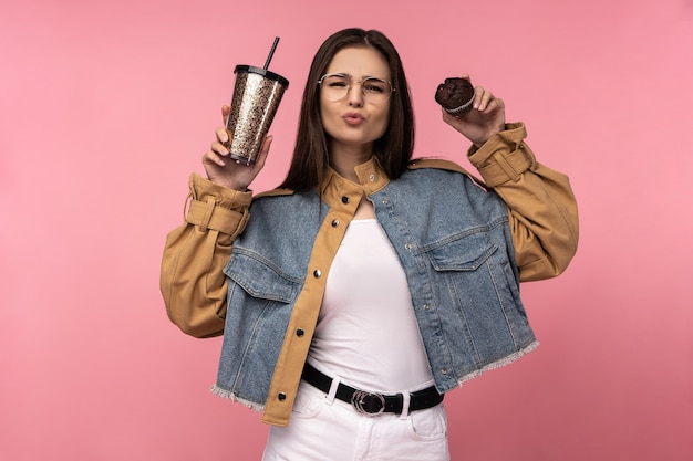 Photo of attractive lady holds tea coffee muffins happy shows love, wears casual jeans jacket white undershirt isolated pink color background.