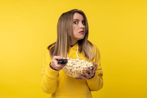 Photo of attractive lady holds tv remote and plate of popcorn, with disgruntled face tries to turn a channel. Wears casual yellow hoody, isolated yellow color background.