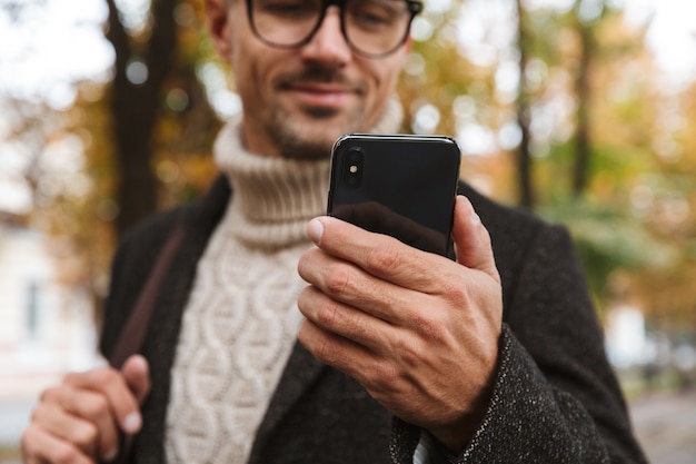 Foto di uomo attraente anni '30 che indossa vestiti caldi che cammina all'aperto attraverso il parco d'autunno e utilizza lo smartphone