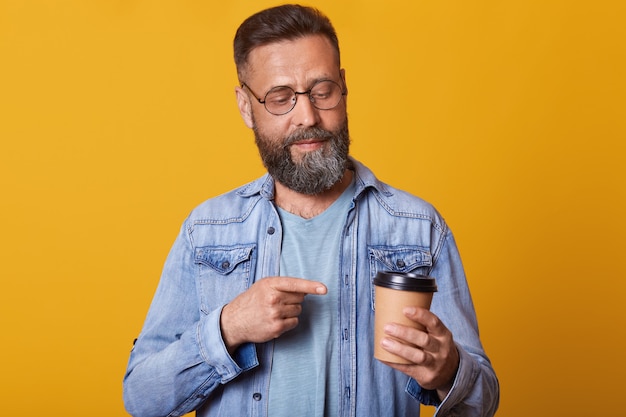 Foto di uomo attraente che punta sulla tazza di caffè o tè, guardando la tazza disponsable, in posa isolato su muro giallo