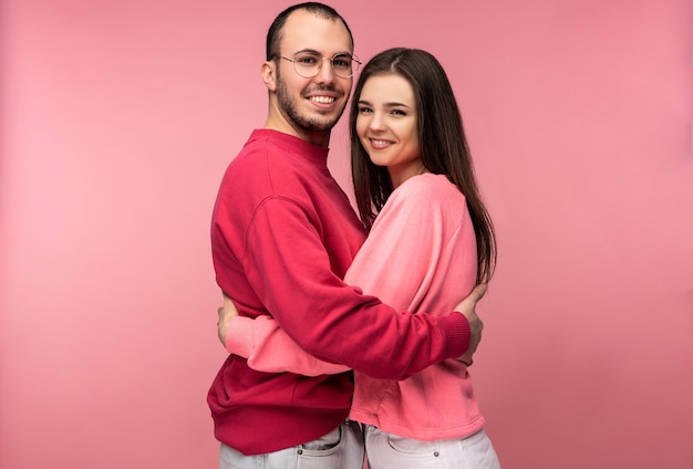Photo of attractive man wih beard in red clothing and woman in pink hug each other and smile. Couple looks happy, isolated over pink background.