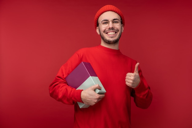 Photo of attractive man with beard in glasses and red clothing. Male got gift and gives thumb up, isolated over red background.