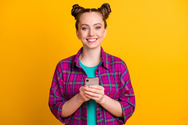 Photo of attractive pretty funny teen lady two buns hold telephone hands charming smile good mood blogger wear casual plaid magenta shirt isolated yellow bright vivid color background