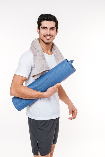 Photo of attractive sportsman with towel in gym standing over white wall.