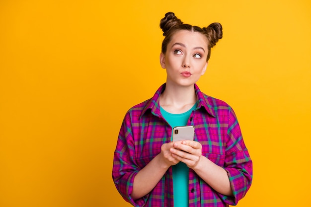 Photo of attractive teen lady two buns hold telephone hands blogger look up dreamy empty space write creative post wear casual plaid magenta shirt isolated yellow bright color background