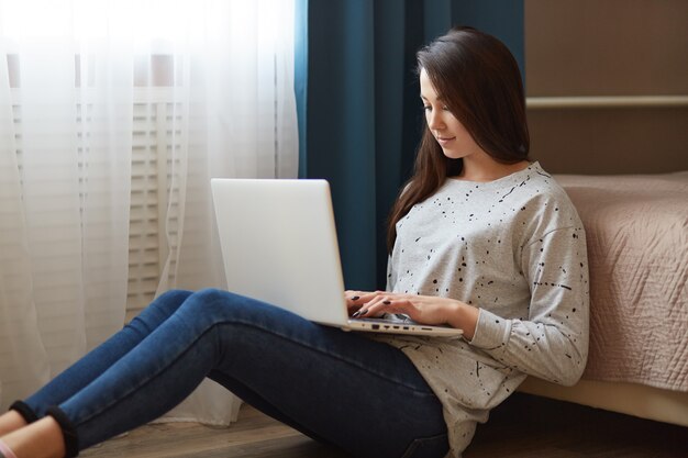 Photo of attractive woman dressed in stylish sweater and jeans, keeps laptop computer on knees, keyboards information, focused at monitor