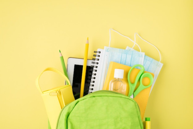 Above photo of backpack notepad pen pencil tablet pencilcase mask and sanitizer isolated on the yellow background